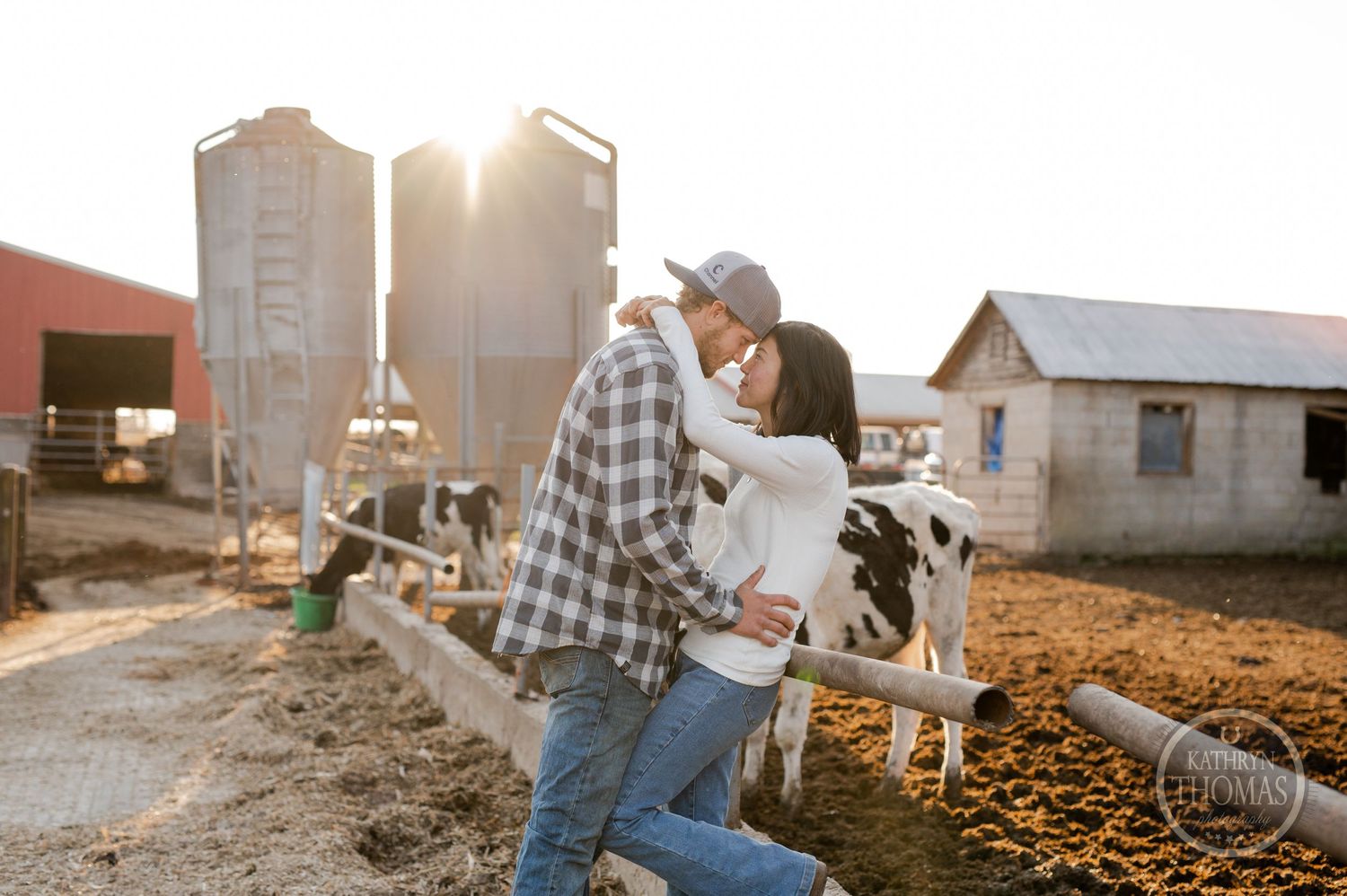 A couple embraces in front of grain silos during golden hour on a farm.