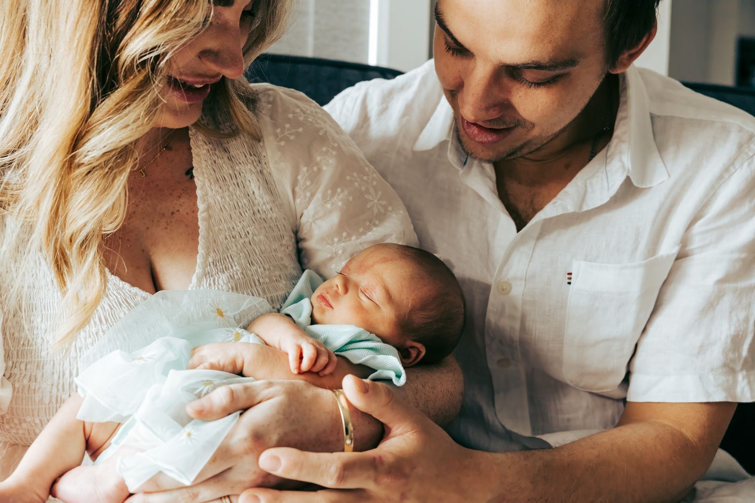 Parents tenderly cradling their newborn baby dressed in light blue while sitting together.
