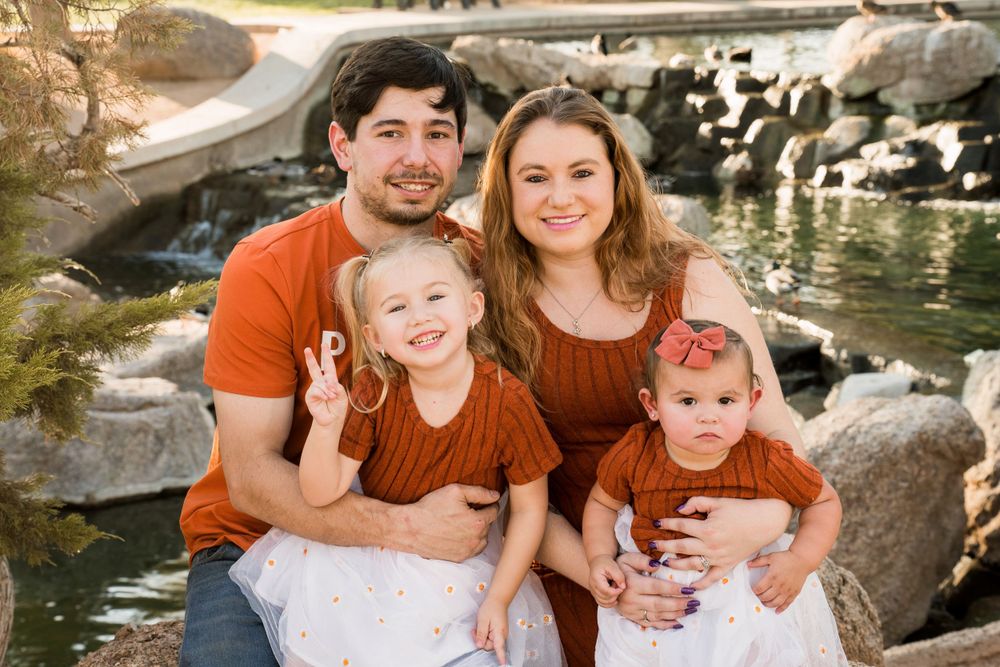 Family of four wearing coordinating rust-colored outfits poses together by a rock pond in outdoor portrait session.