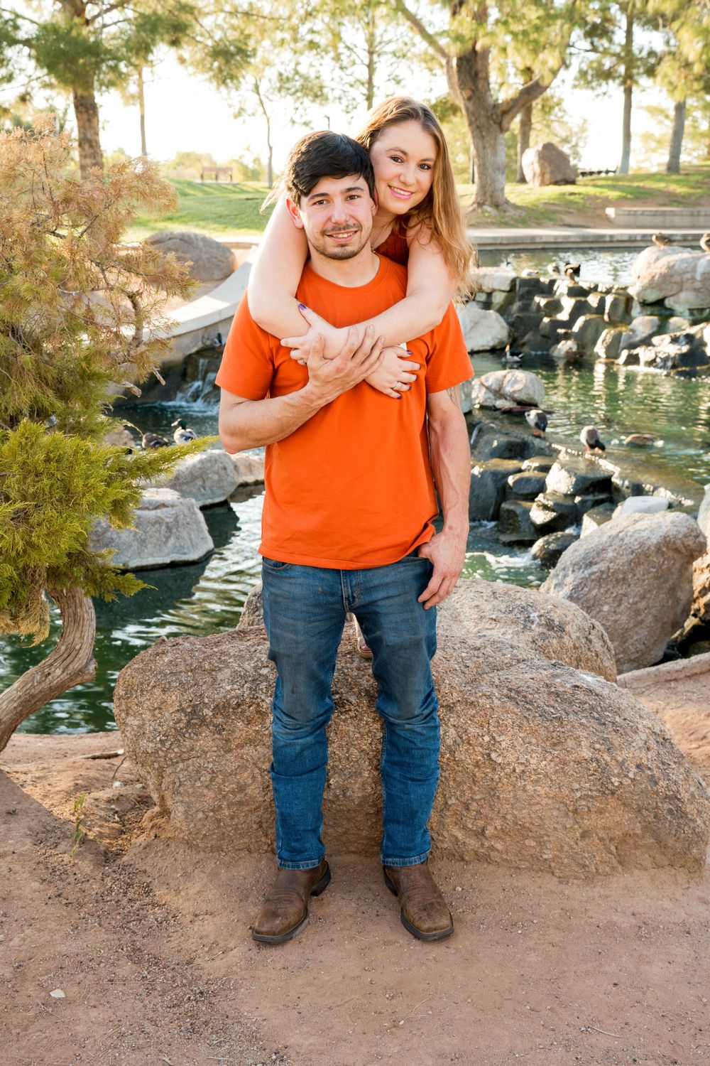 Couple embraces while standing on rocky terrain near a decorative pond during outdoor photo shoot.