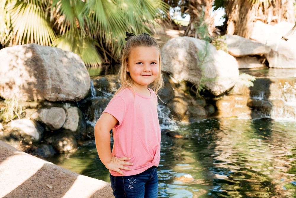 Child in pink shirt poses confidently by decorative rock pond with palm trees in background.