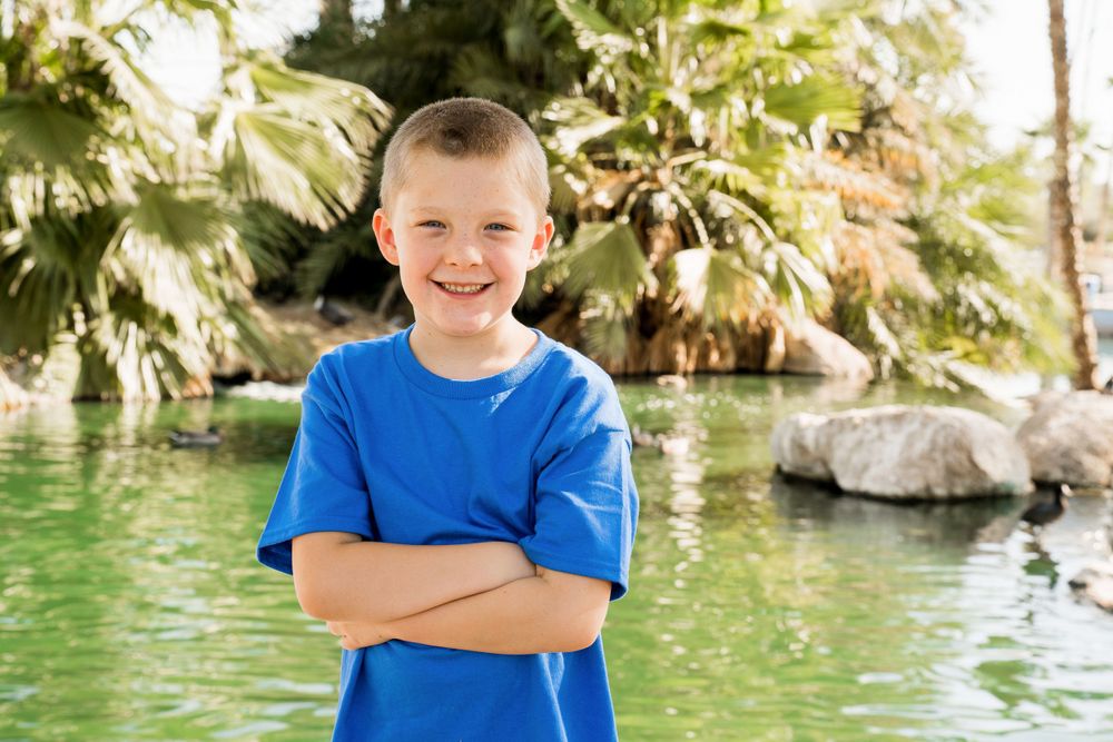 Young child in bright blue t-shirt poses with crossed arms by tropical water feature.