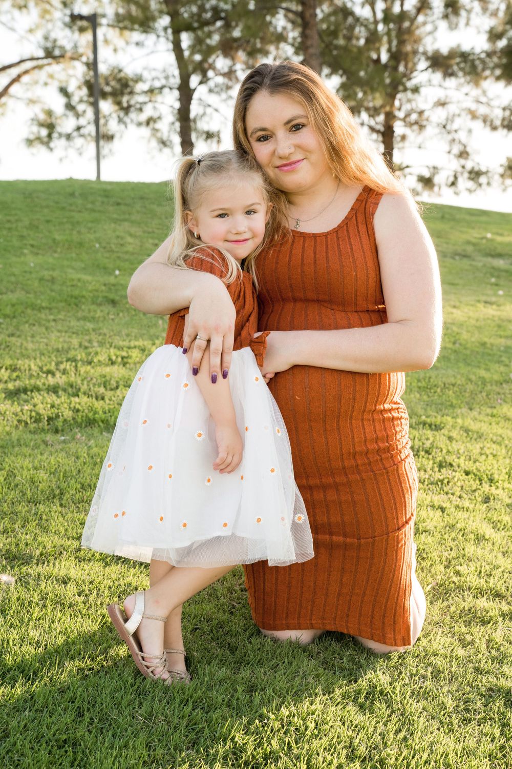 Mother and daughter in matching rust dresses pose on green grass during sunset photo session.