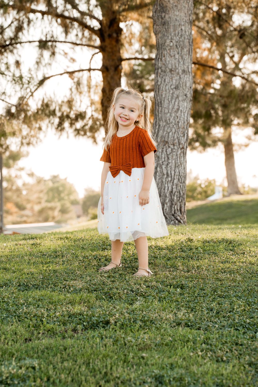 A small child stands on grass wearing an orange and white dress near pine trees.