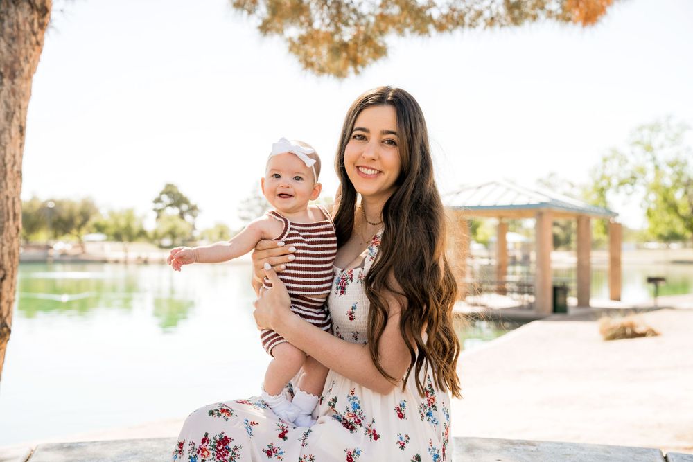 Mother holding baby poses outdoors near water feature on sunny day.