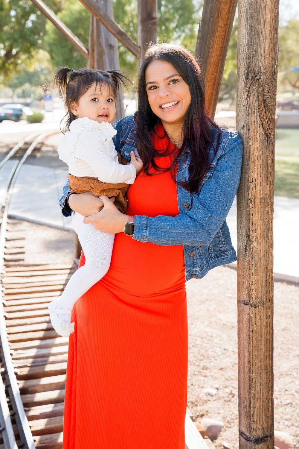 A mother and child pose together near a wooden structure, with her wearing a red dress and denim jacket.