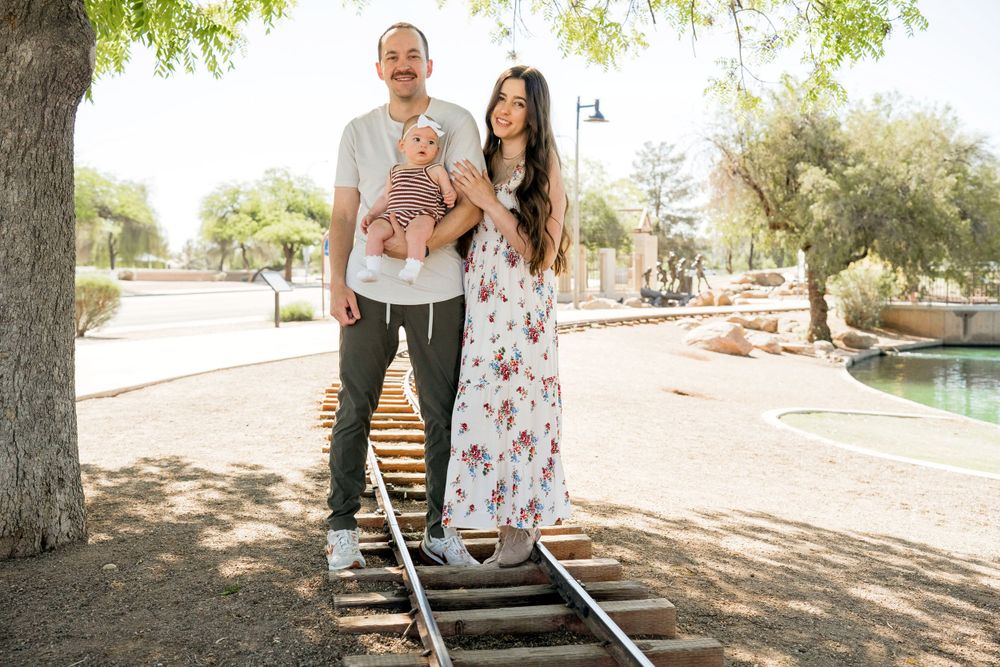 Couple poses with baby while standing on decorative train tracks in park setting.