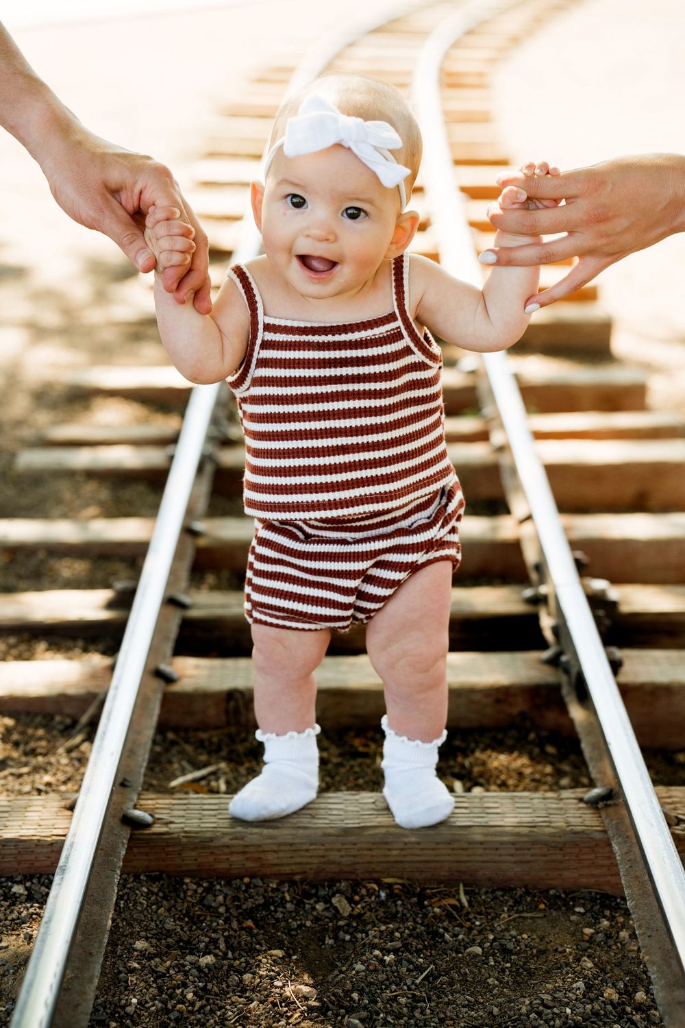 Baby in striped romper stands on train tracks while holding parents' hands.
