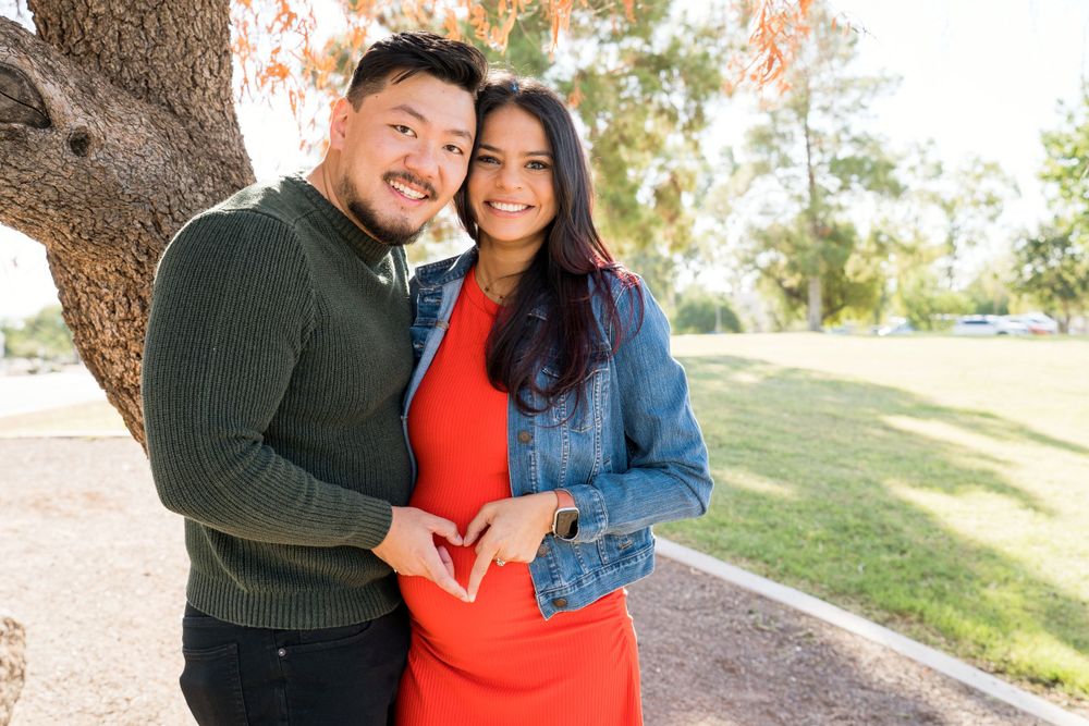 A couple poses together outdoors, with the woman wearing a red dress and denim jacket.