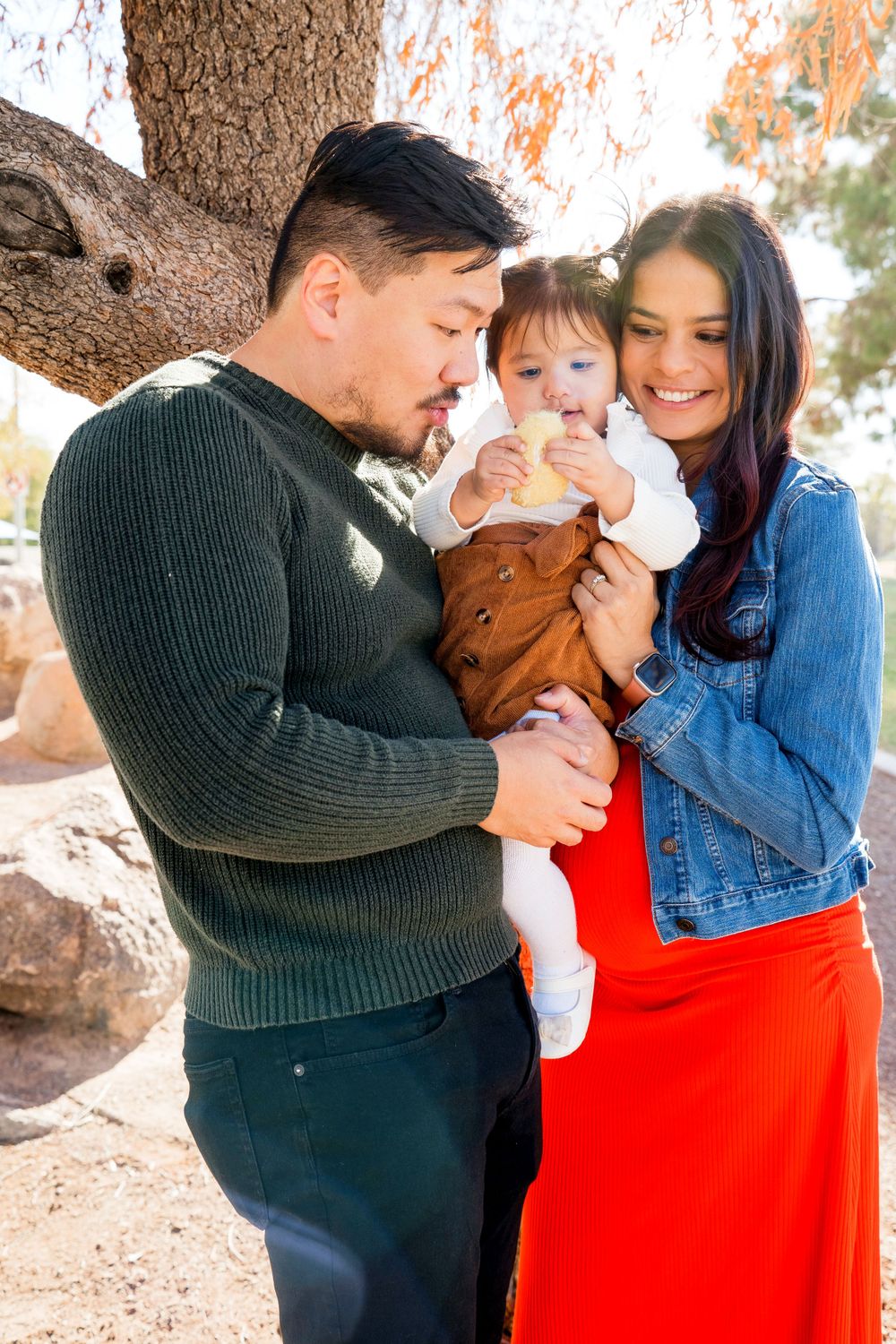 A couple shares a tender moment with their baby outdoors under a tree on a sunny day.