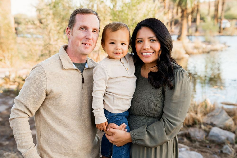 Family of three posing outdoors for a portrait near a waterfront with palm trees.