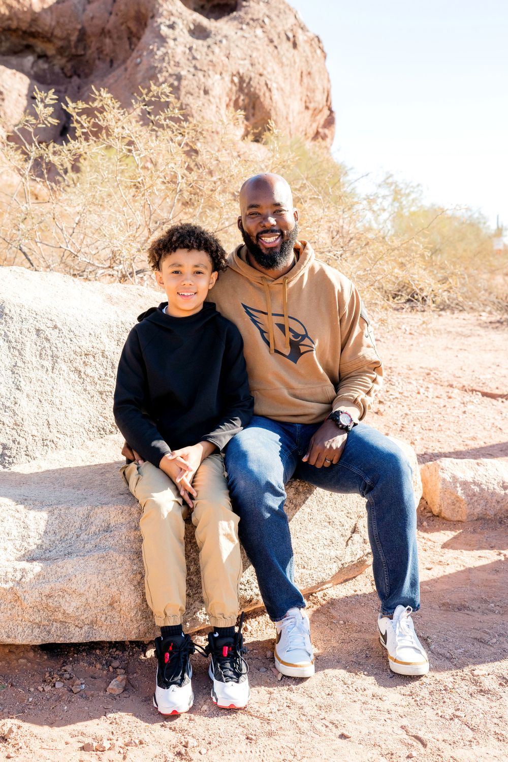 A father and son sit together on rocky terrain wearing casual clothing.