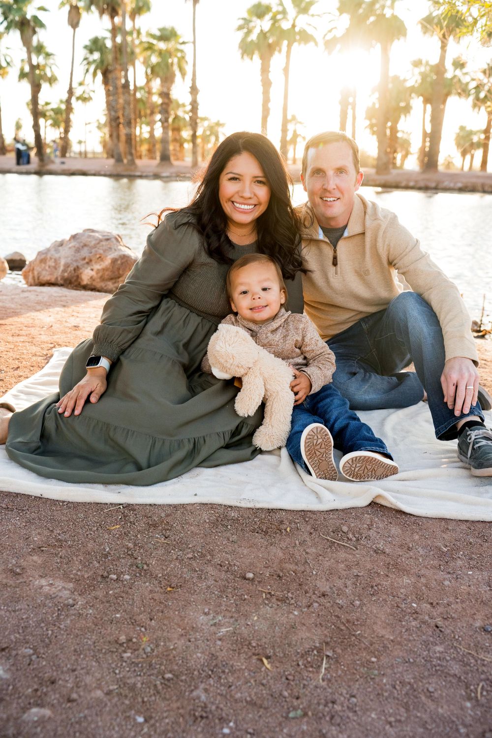 Family sitting on a blanket by a lake during golden hour with palm trees in the background.