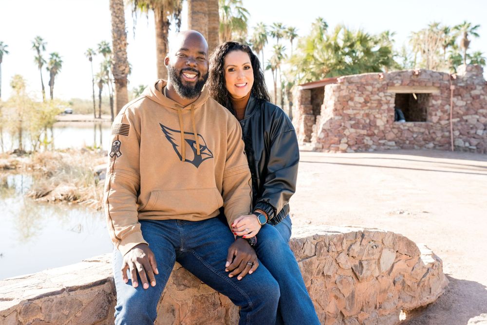 Couple sitting together on rocks wearing casual clothing with palm trees in background.