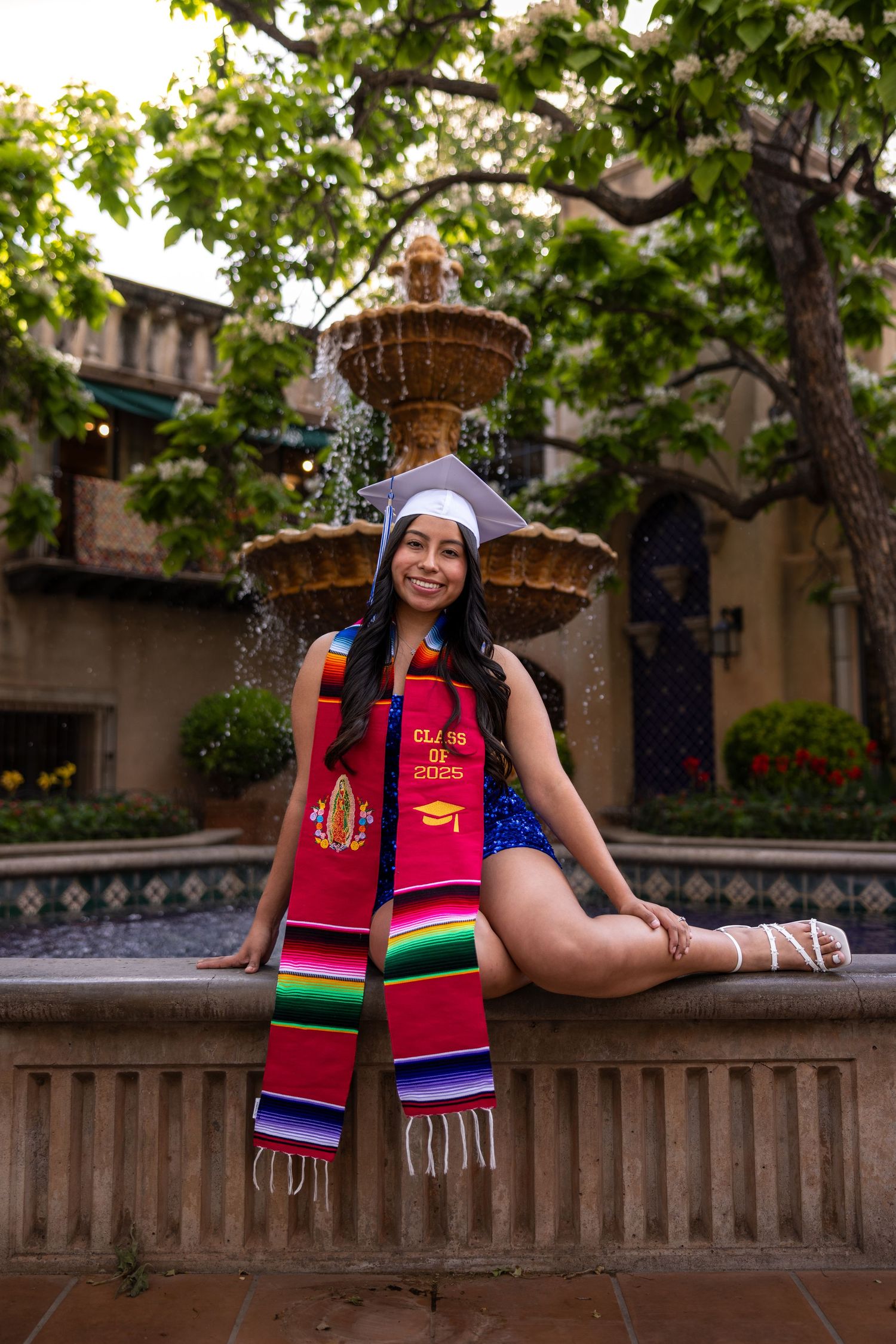 Graduate poses in cap and colorful sash by ornate fountain in Spanish-style courtyard.