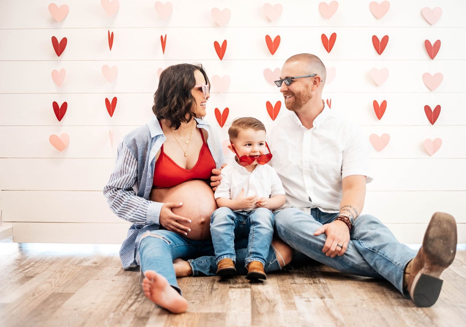 Pregnant family sitting on floor with heart decorations celebrating Valentines Day maternity session.
