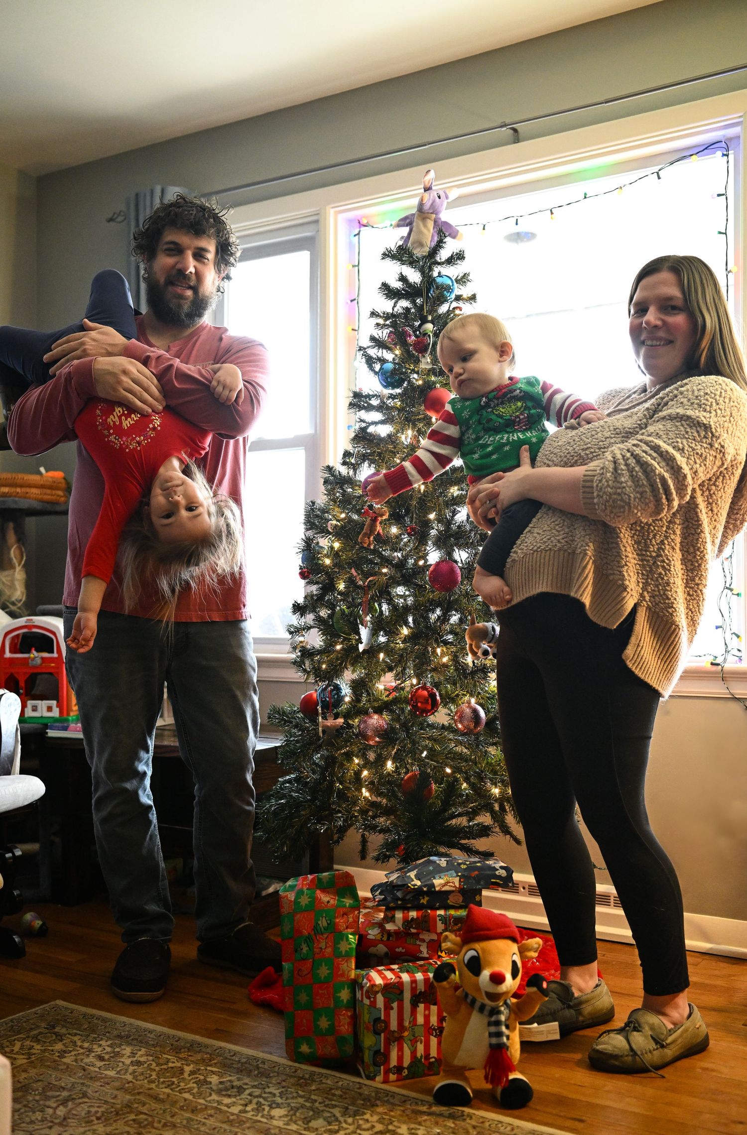 A man and a woman in front of a Christmas tree in their home, the man holding their daughter and the woman holding their son. They are happy