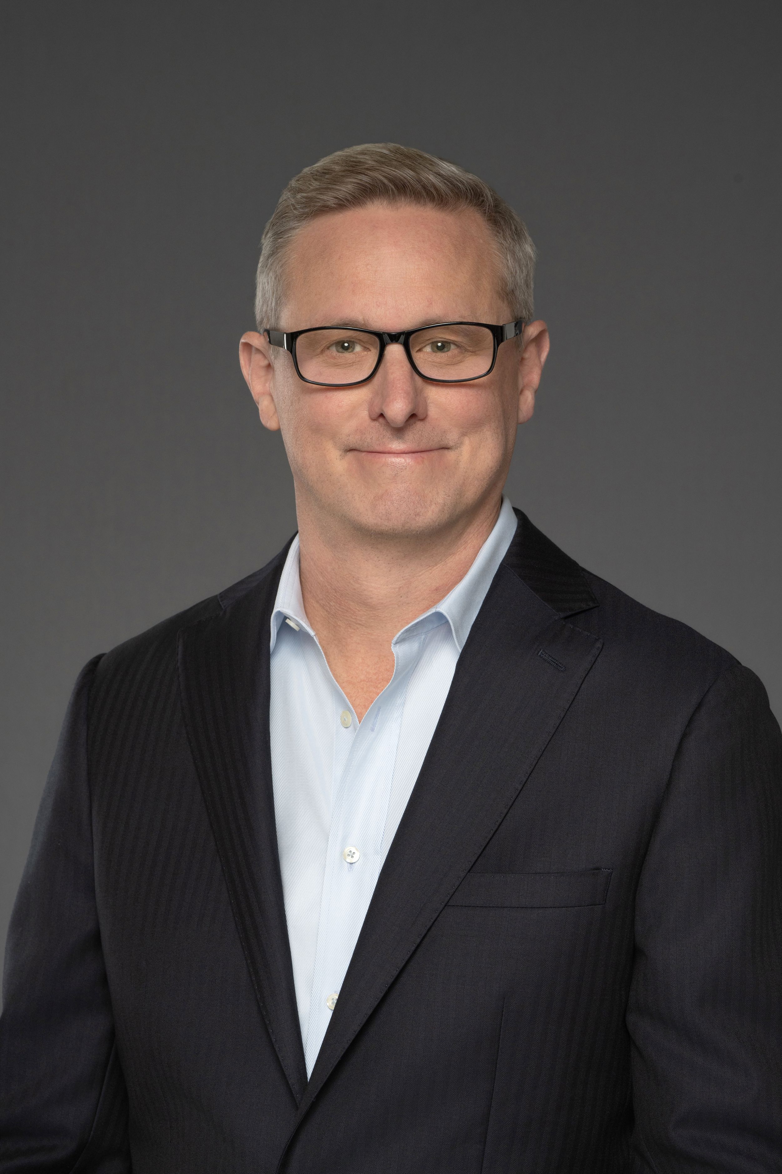 Middle-aged man with gray hair wearing glasses and a dark pinstripe blazer against gray background.