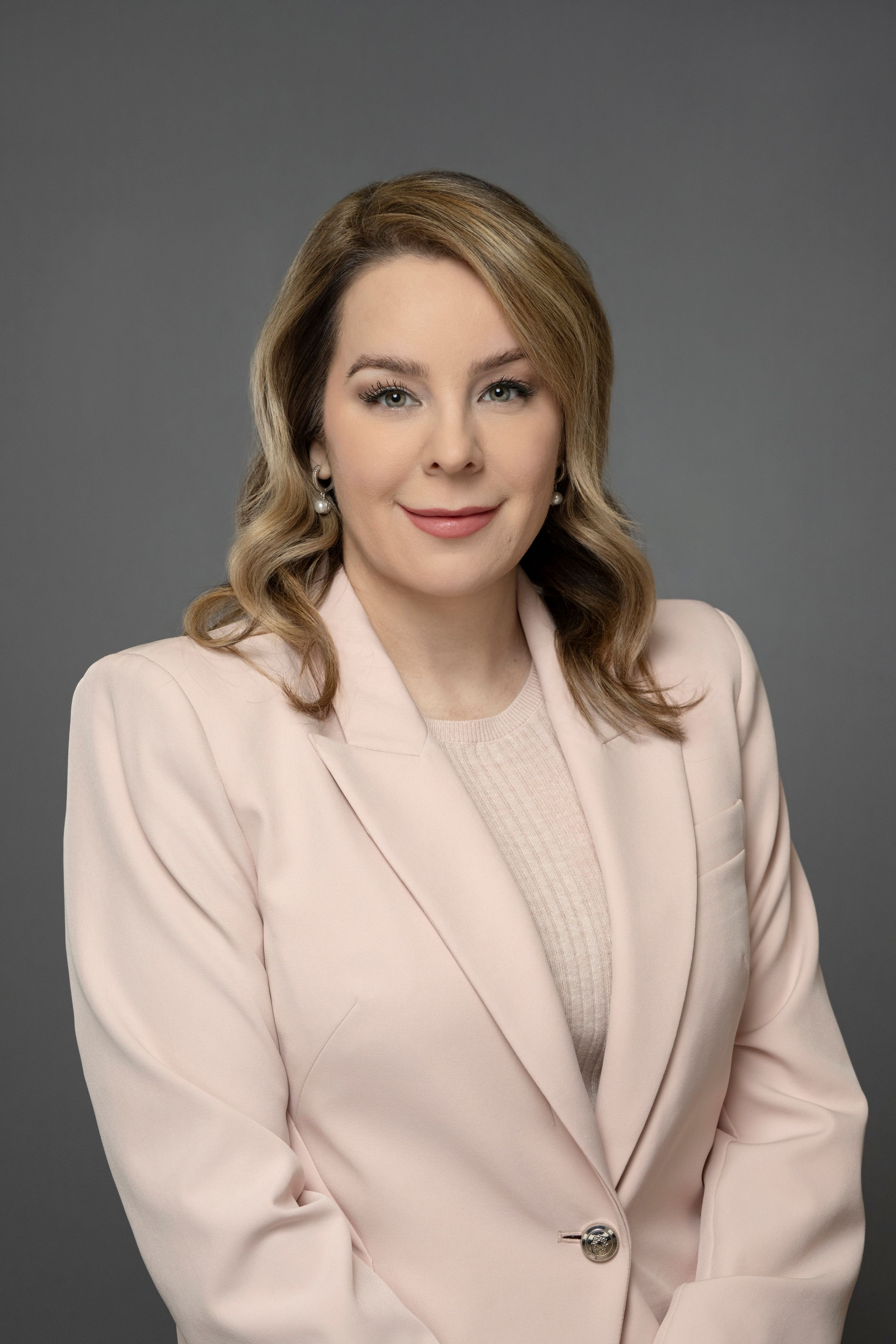 Blonde woman in a pink blazer smiling professionally against a neutral gray background.