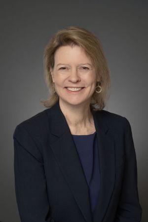 Professional woman with blonde hair smiles in navy blazer for corporate portrait on gray background.