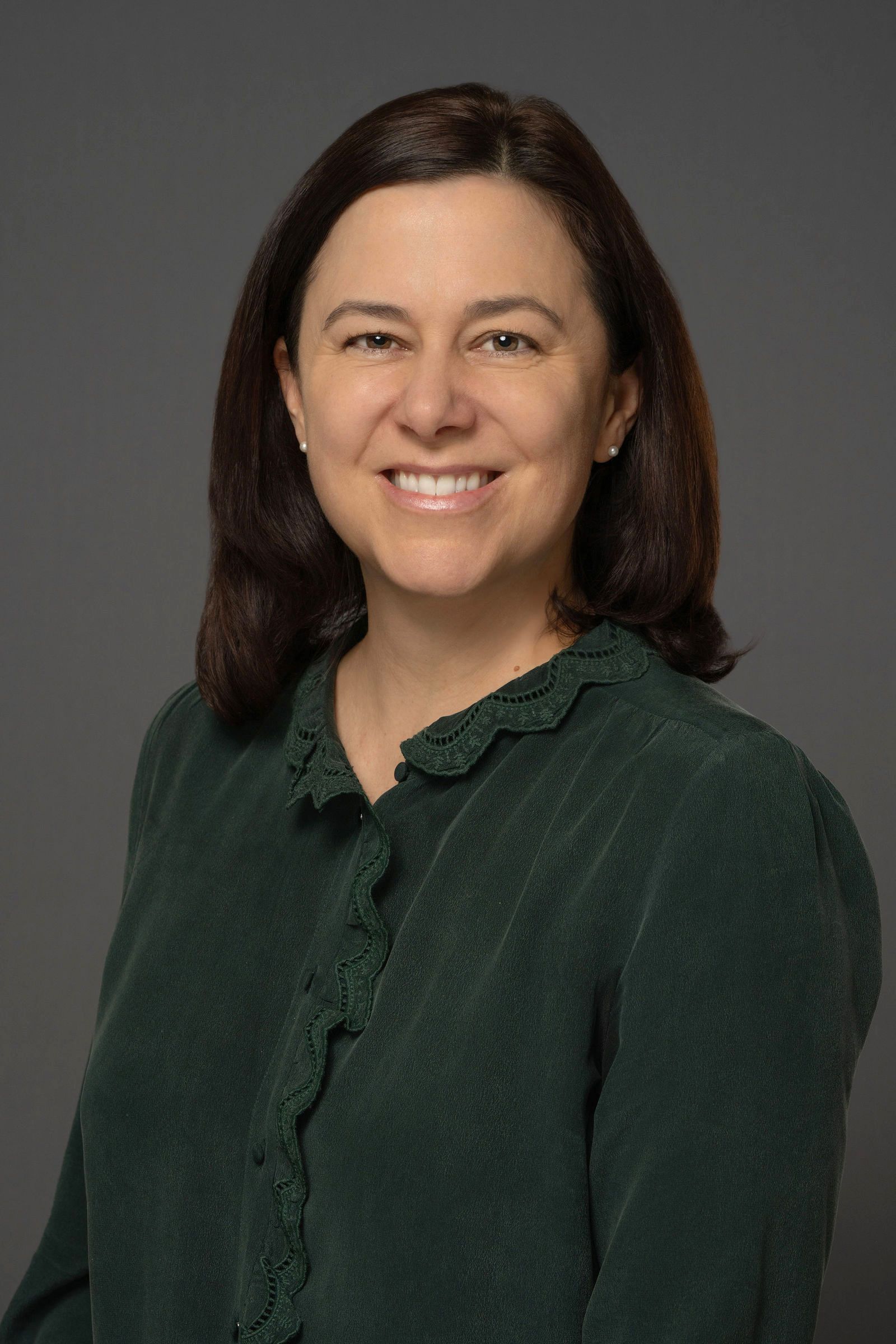 Professional woman with dark hair smiles in green blouse for corporate headshot on gray background.
