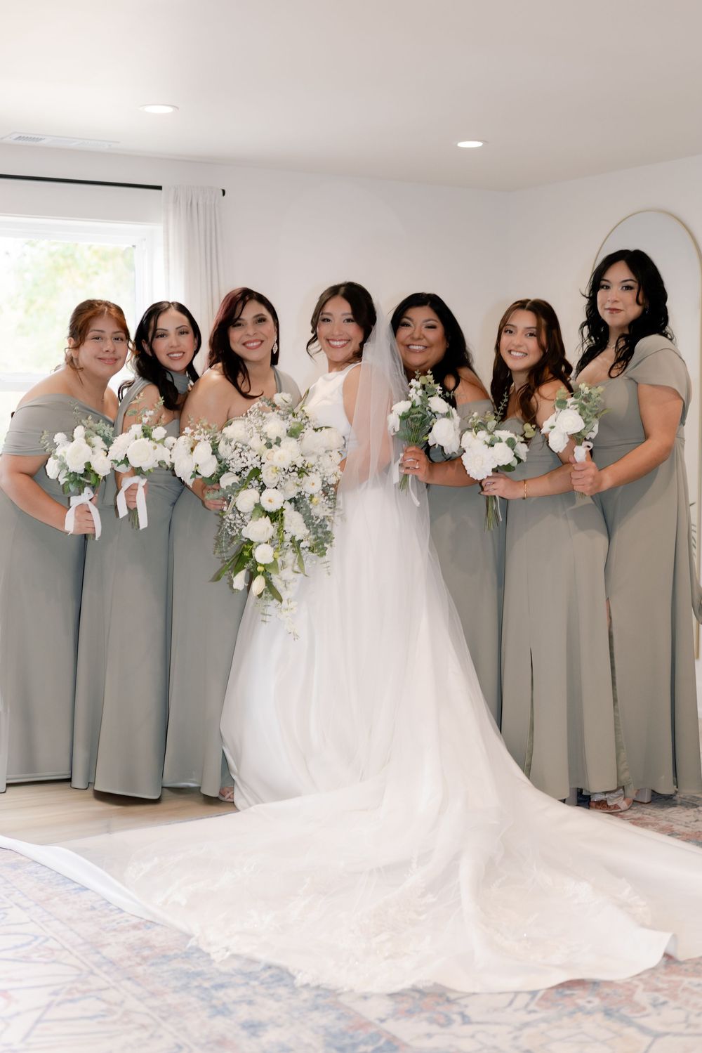 Wedding party in sage green bridesmaid dresses pose with bouquets of white flowers against a light interior wall.