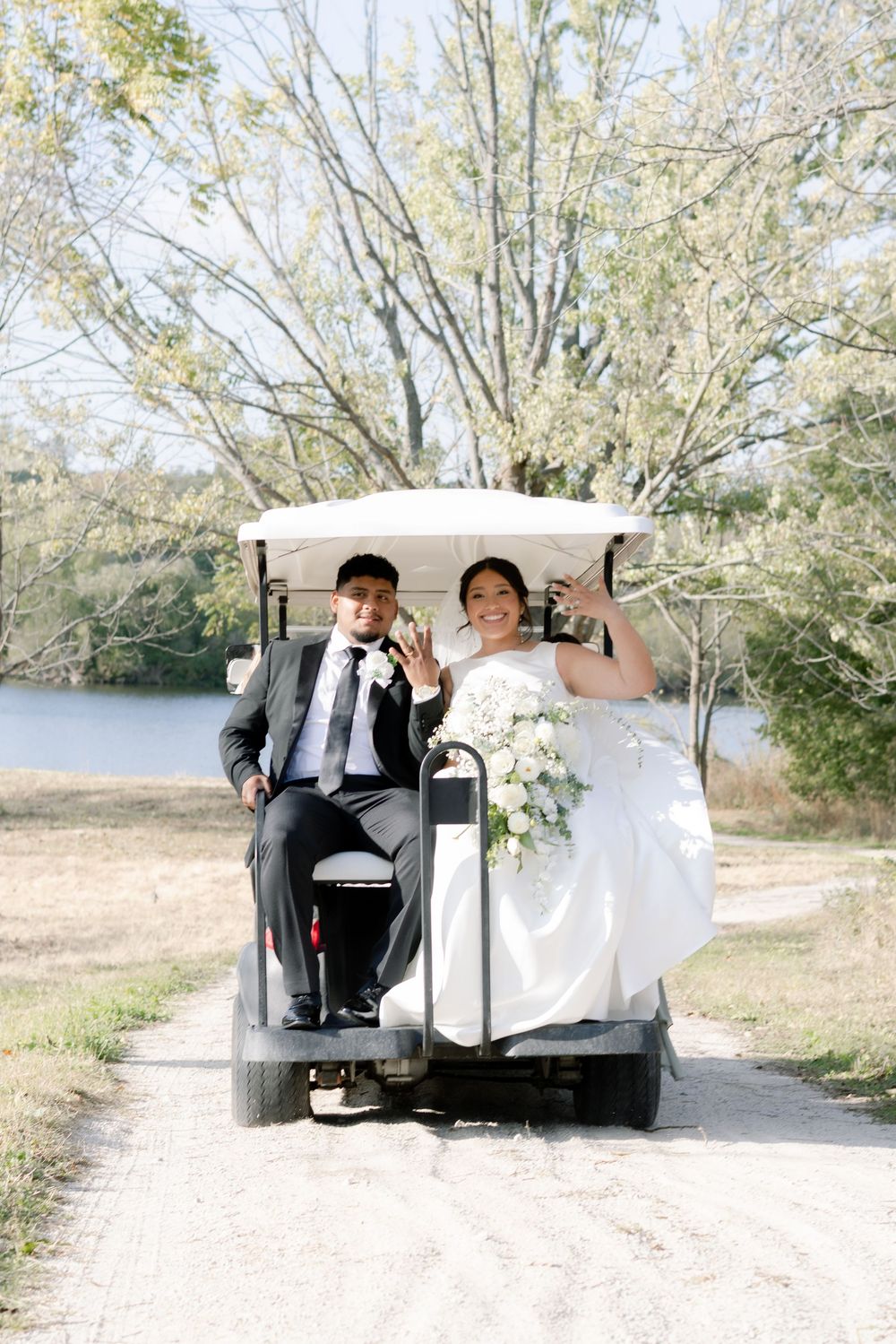 A wedding couple drives away in a golf cart on a scenic path by a lake during their celebration.