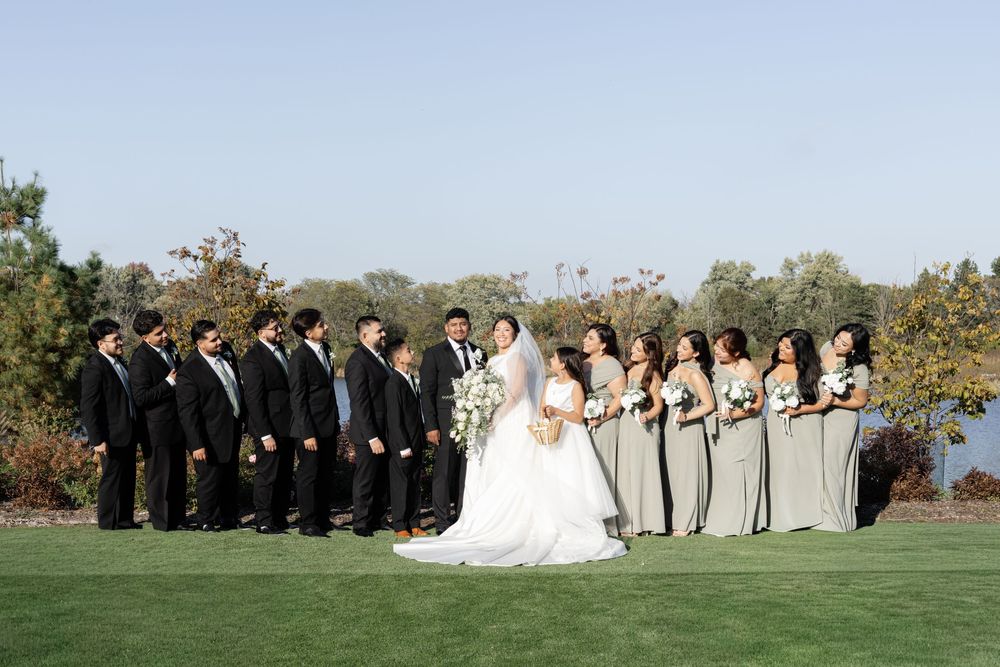 Wedding party in formal attire poses together outdoors in a line against a natural autumn backdrop.
