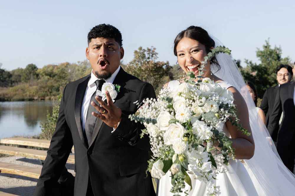Wedding party celebrates joyfully outdoors near a lake with elegant white floral bouquets and formal attire.