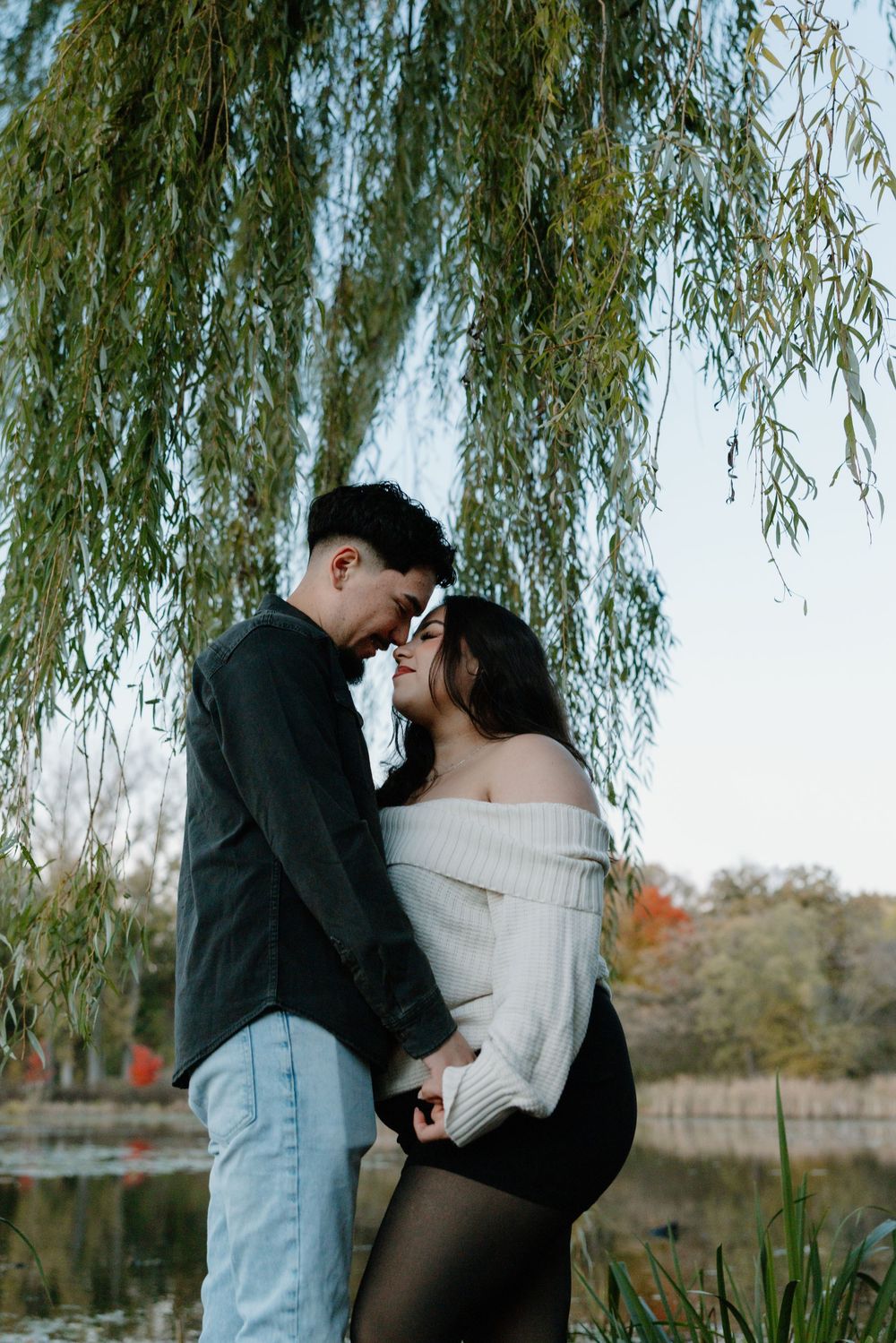 A romantic couple shares an intimate moment under a weeping willow tree beside a tranquil pond.