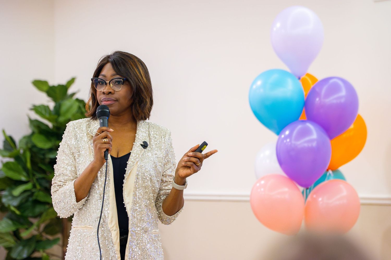 A professional speaker gestures while presenting at an event with colorful balloons and plants in the background.