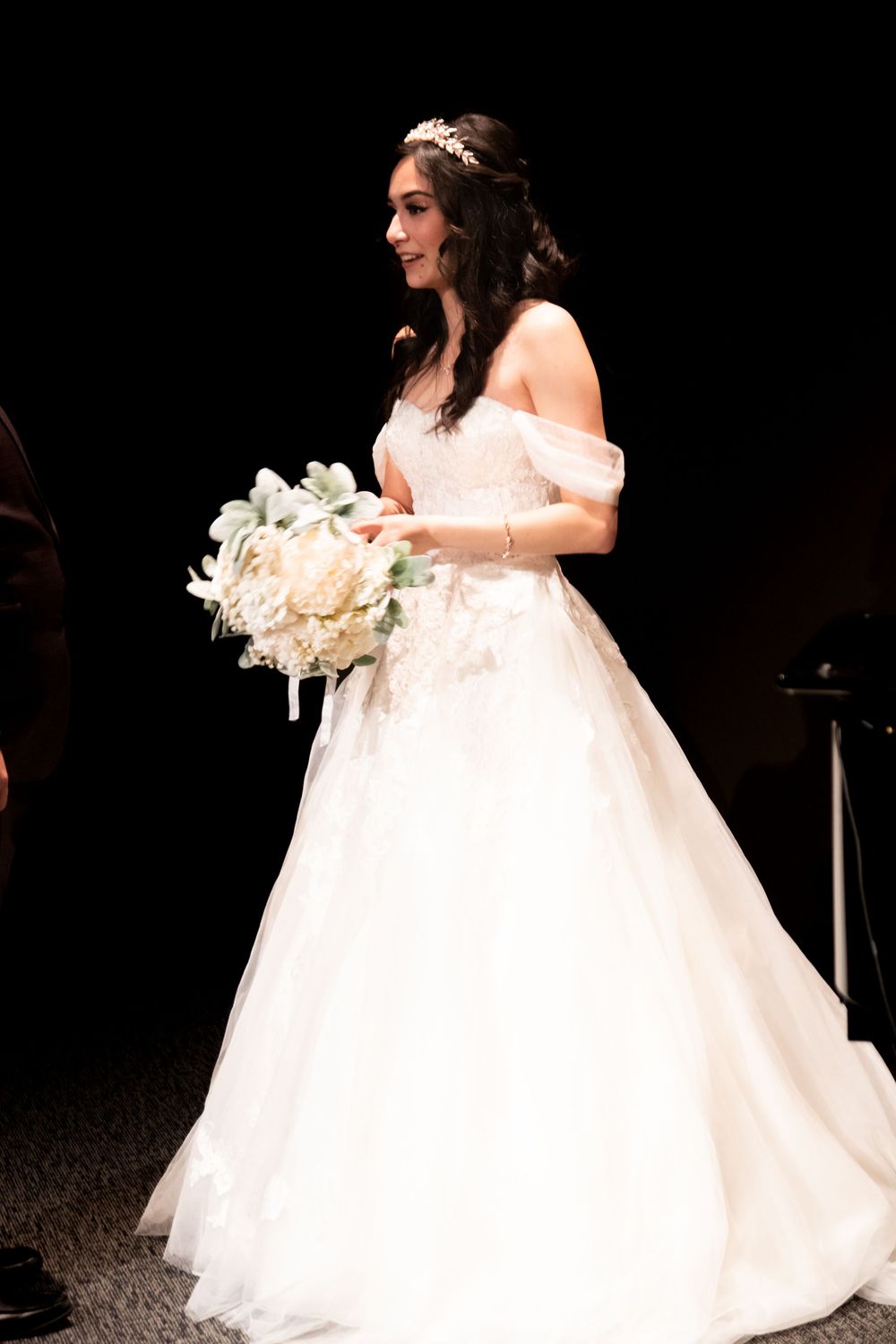 Smiling bride in off-shoulder white ball gown holding white floral bouquet with tiara at wedding ceremony.
