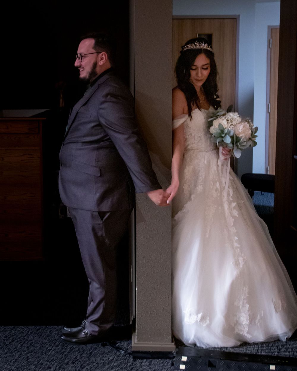 Bride and groom hold hands around corner in first look moment, bride wearing off-shoulder gown and tiara.