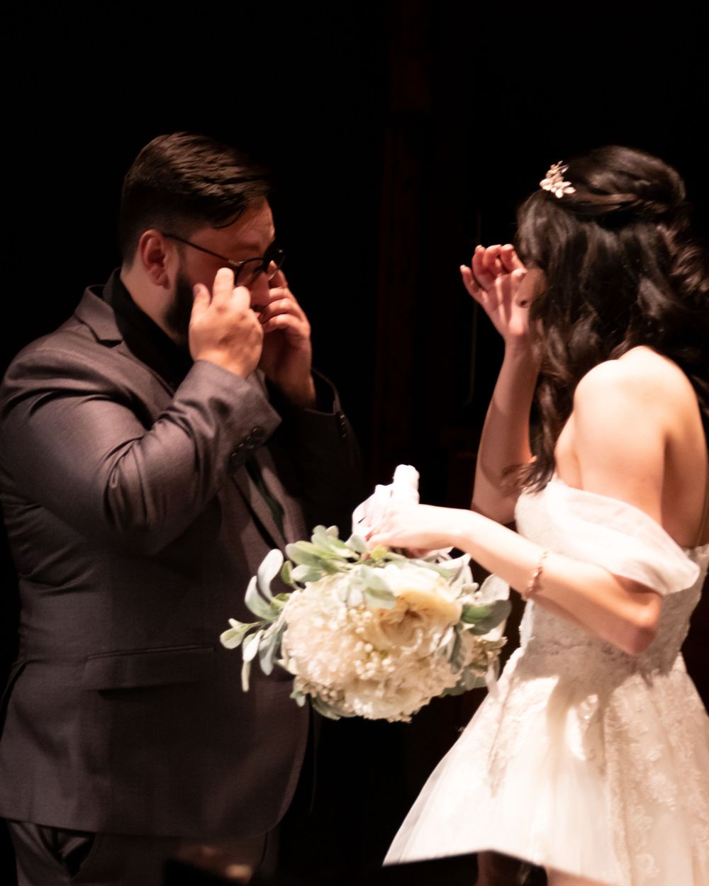 Emotional groom wiping tears while bride holds white bouquet during heartfelt wedding ceremony moment.