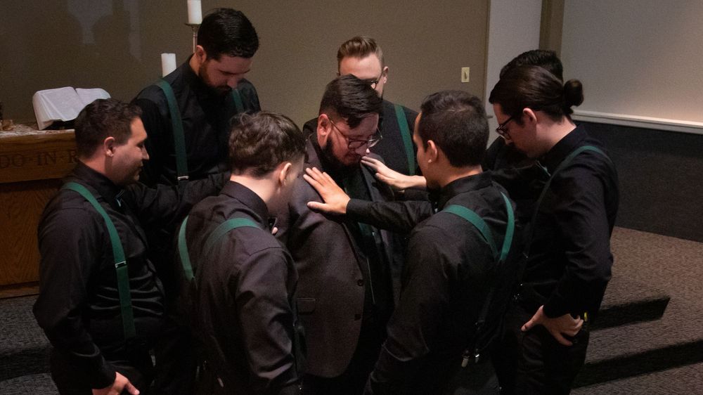 Group of men in black shirts and green suspenders huddle together in prayer inside a meeting room.