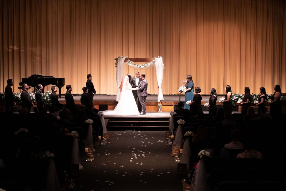 A bride and groom exchange vows at an elegant wedding ceremony on a stage with a floral arch and seated guests.