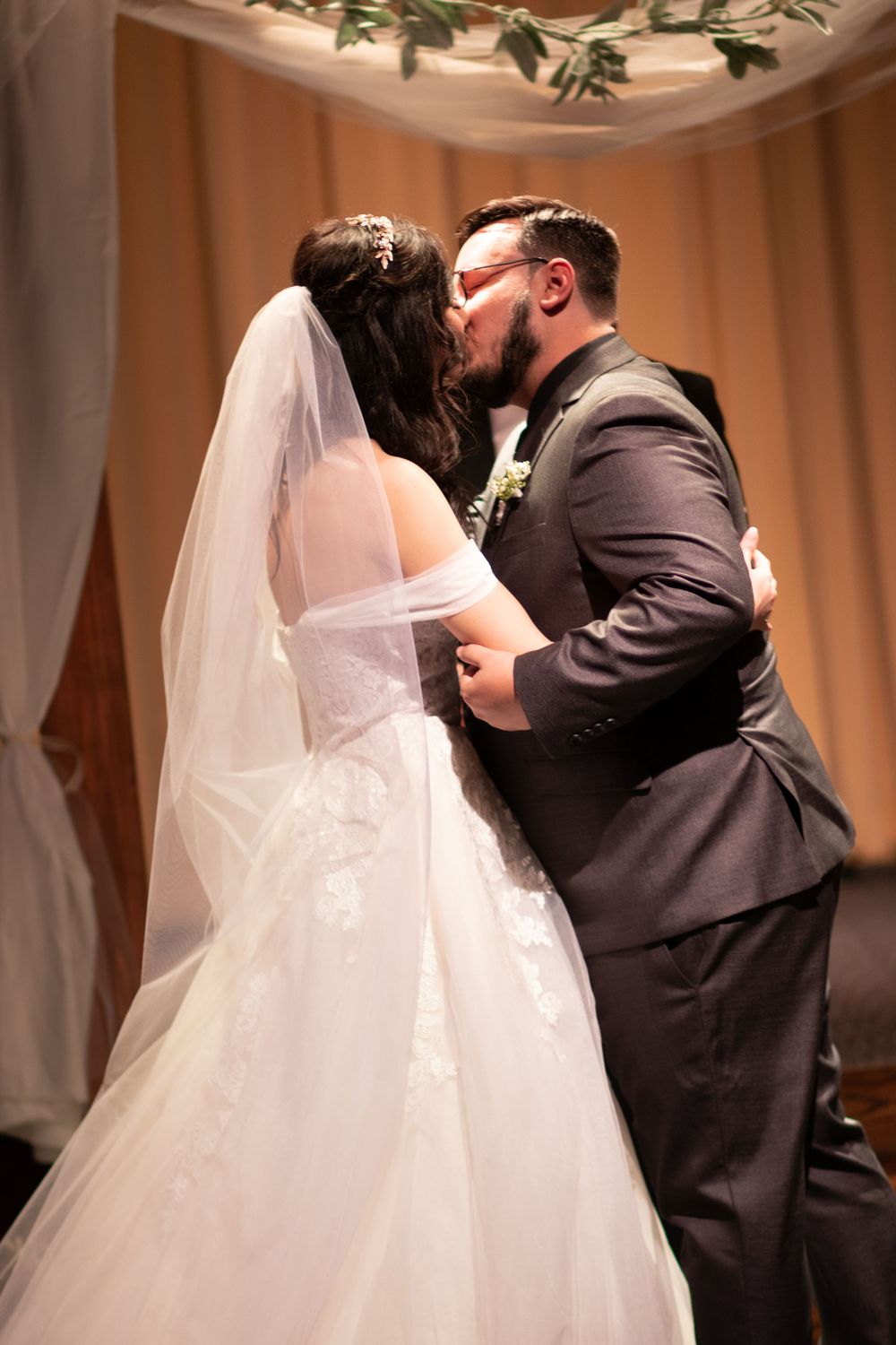 A bride and groom share their first kiss at the wedding altar, with the bride wearing a lace gown and veil.