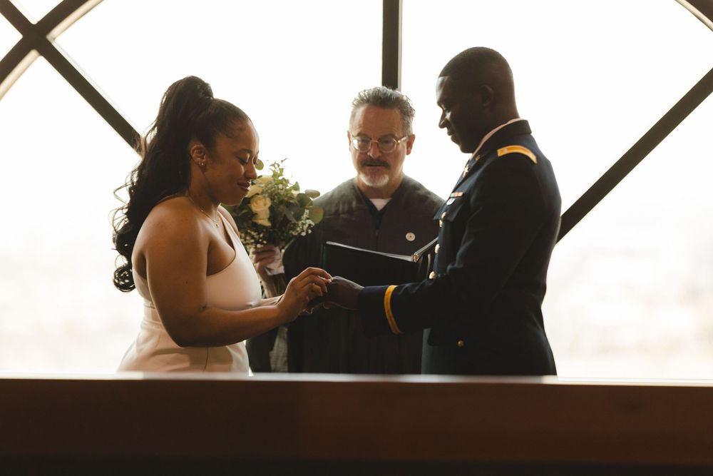 Military groom and bride exchange rings during wedding ceremony with officiant holding bouquet indoors.