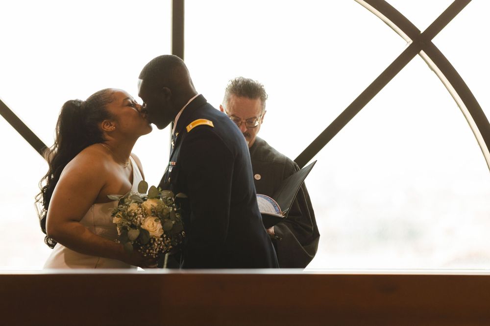 Military groom and bride share a kiss during wedding ceremony with officiant in elegant venue.