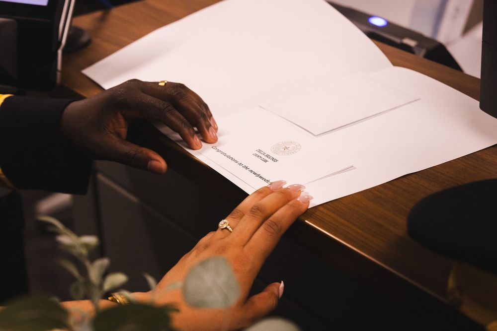 Two hands exchanging documents on a wooden desk, one wearing a diamond ring, in a formal signing ceremony.