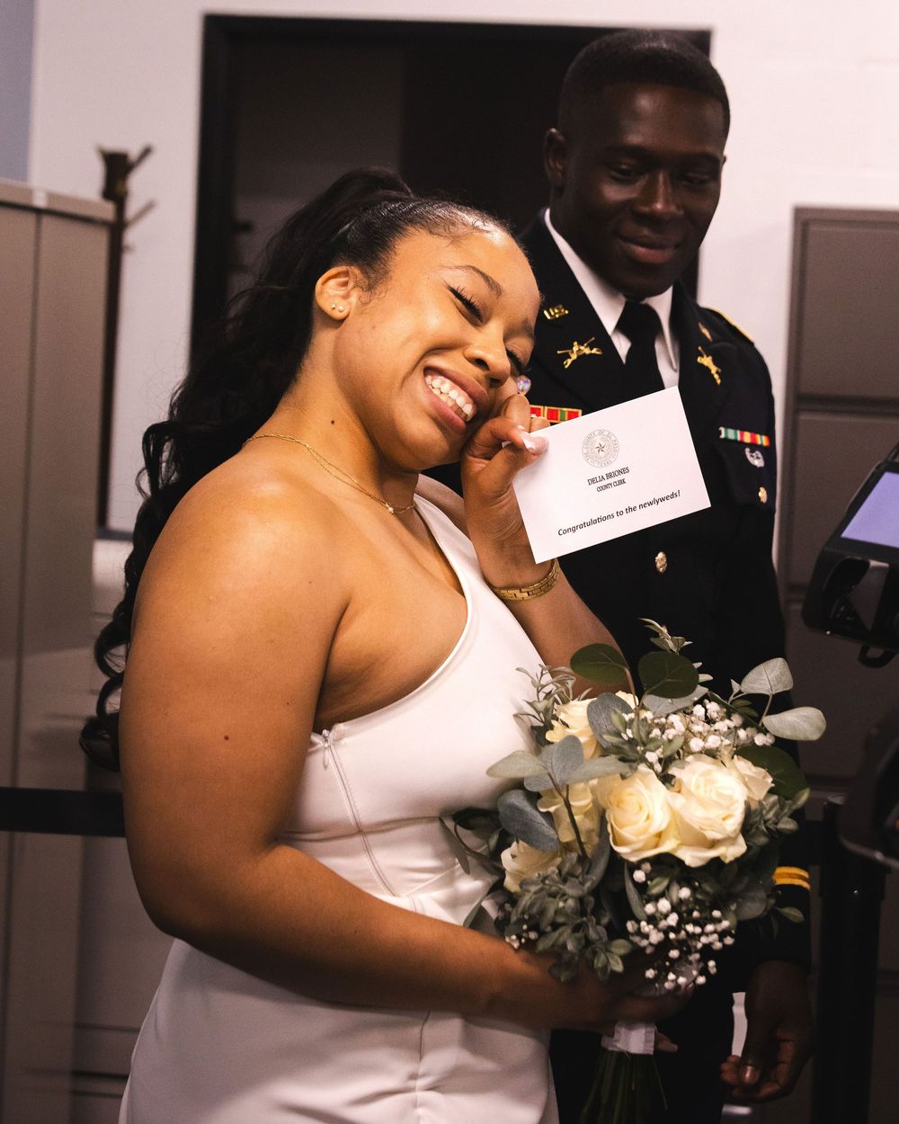 A smiling bride in a white dress holds white roses while a groom in military uniform stands behind her.