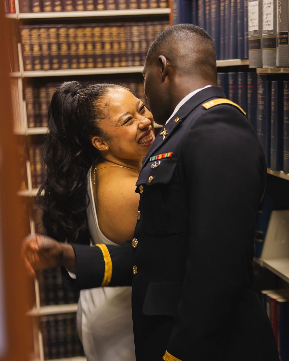 A smiling woman in a white dress embraces a man in US Army dress uniform in a library setting.