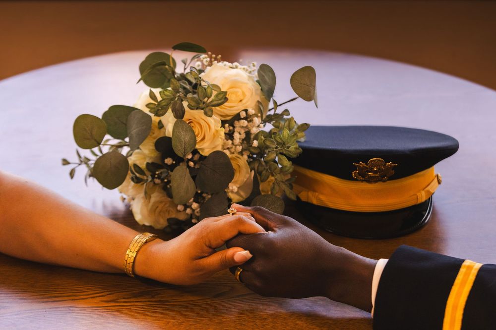 Military officer and partner holding hands on table with white rose bouquet and dress cap at wedding ceremony.