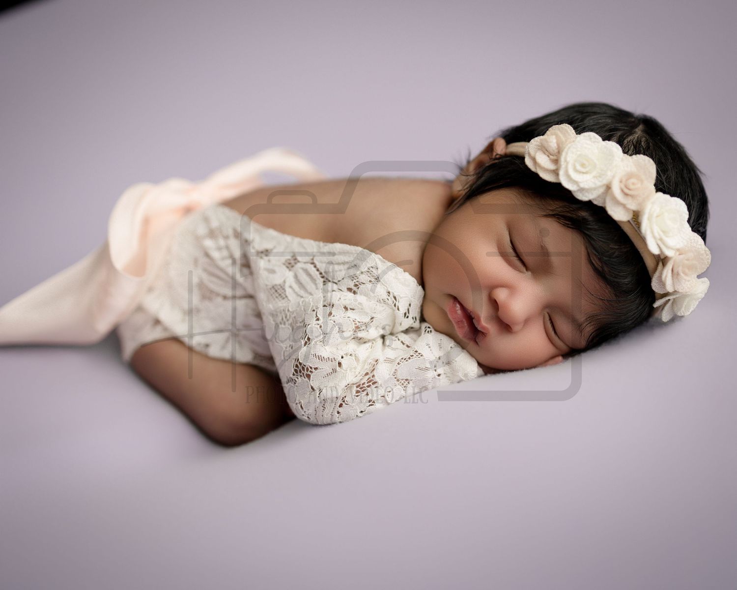 Sleeping newborn wearing white lace dress and floral headband rests peacefully on purple backdrop.