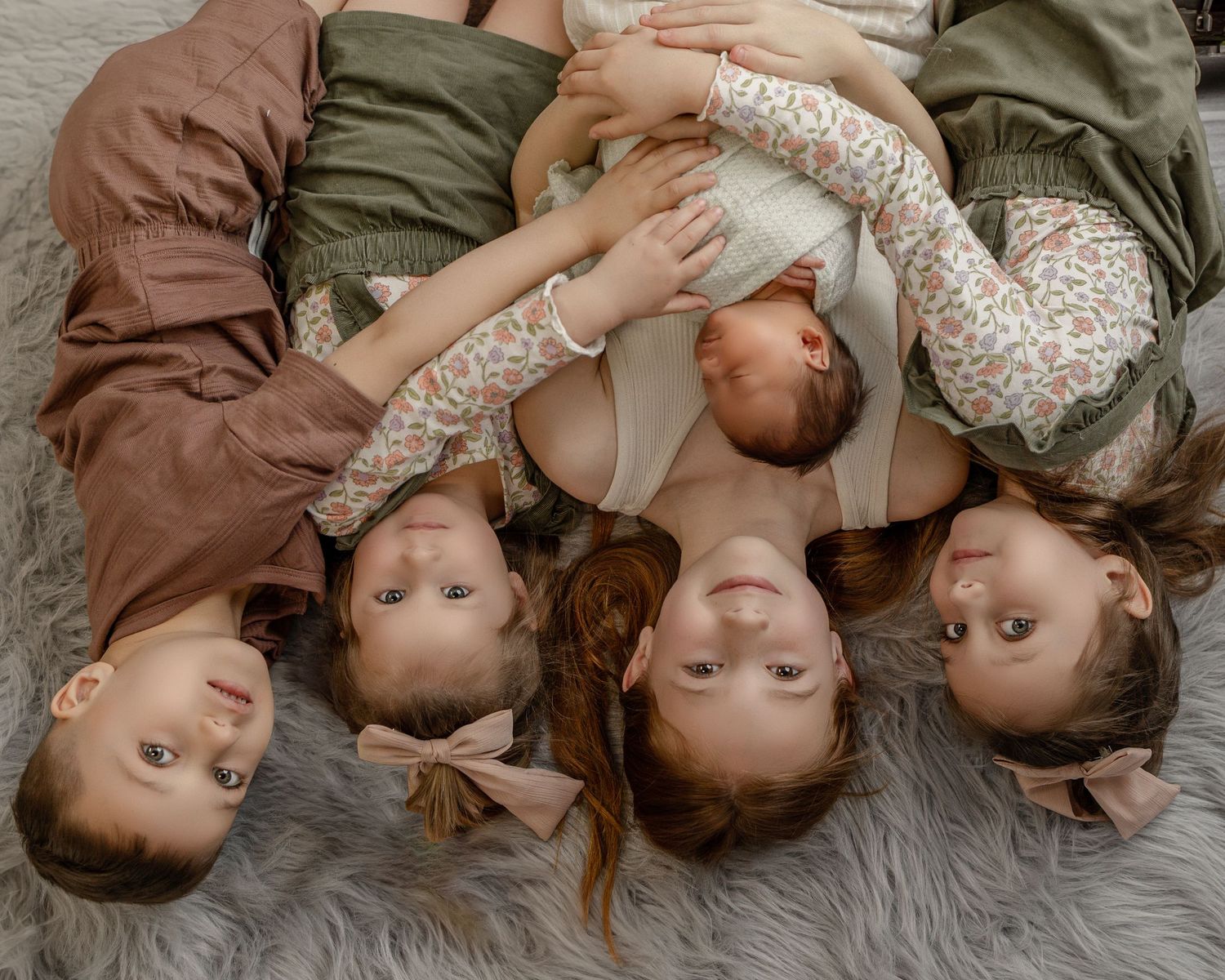Five siblings wearing coordinated outfits lay in a circle on a gray rug looking down at the camera.