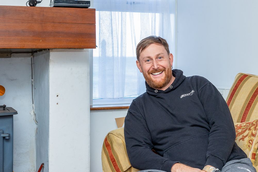 A man with a beard wearing a black jacket and sitting on a chair.