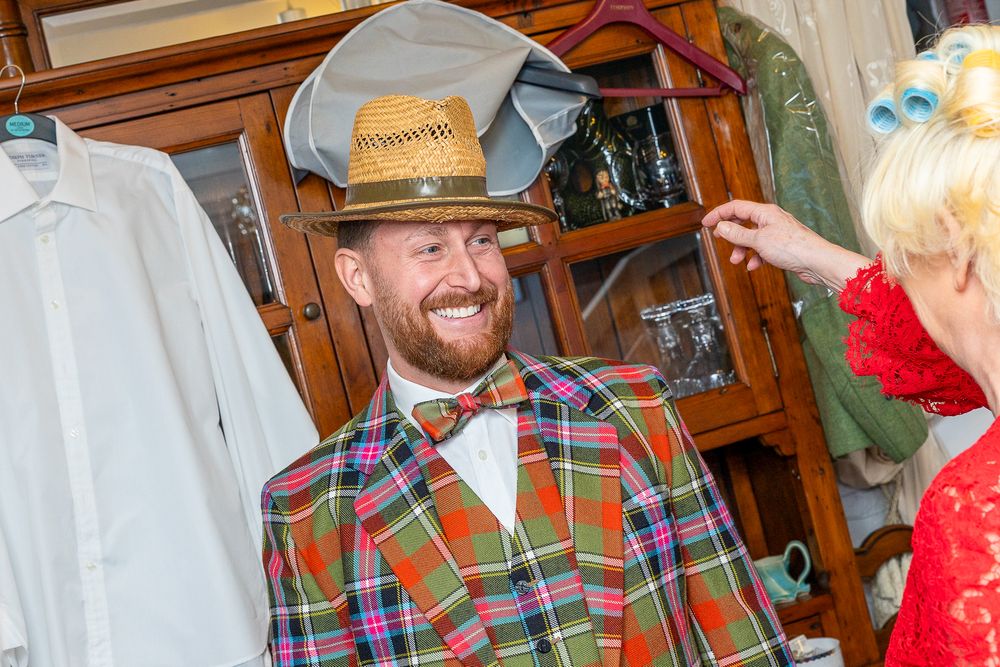 A man with a beard wearing a plaid shirt and a construction worker's hat is smiling at the camera.