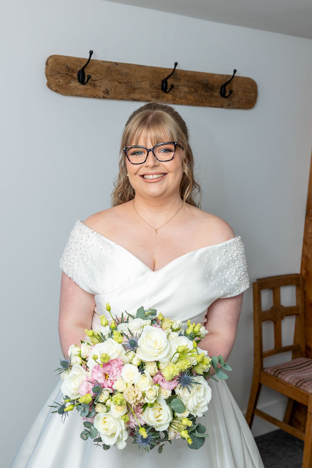 A smiling woman in a white wedding dress holding a bouquet of flowers stands in front of a wooden wall with hooks.