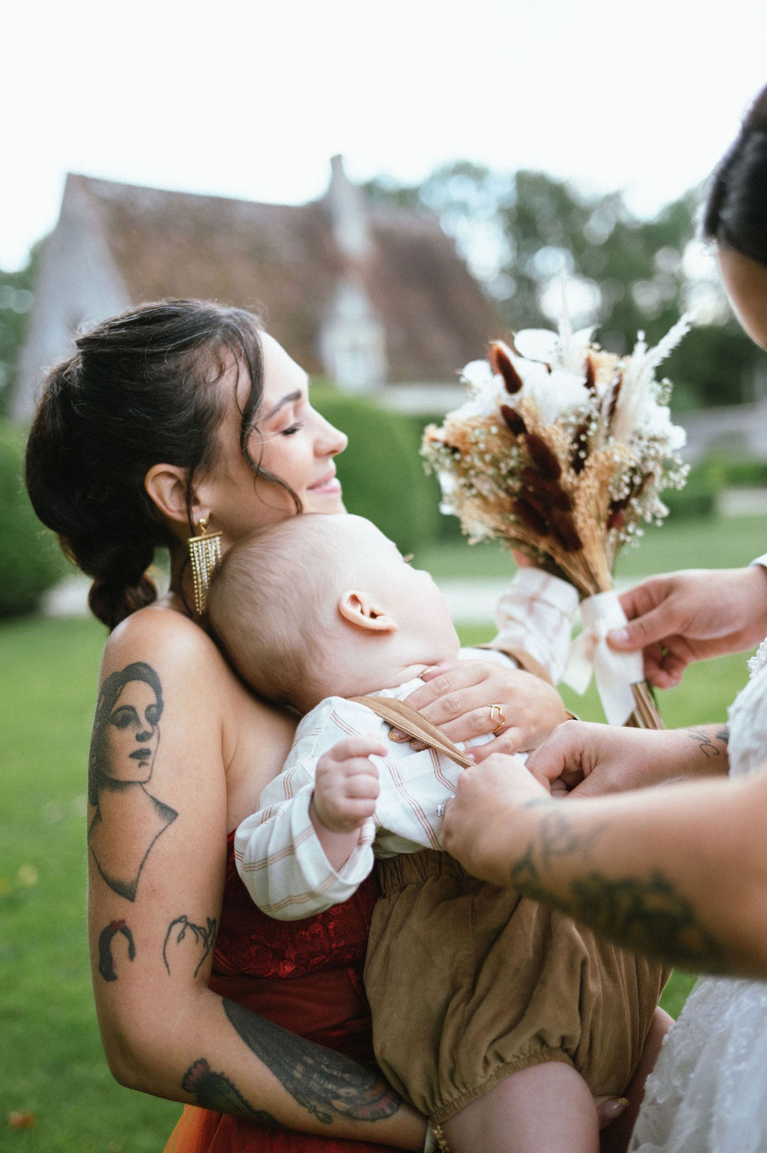Une jeune mère tatouée serre tendrement son bébé dans les bras pendant une cérémonie, une image forte de lien et d’intimité familiale.