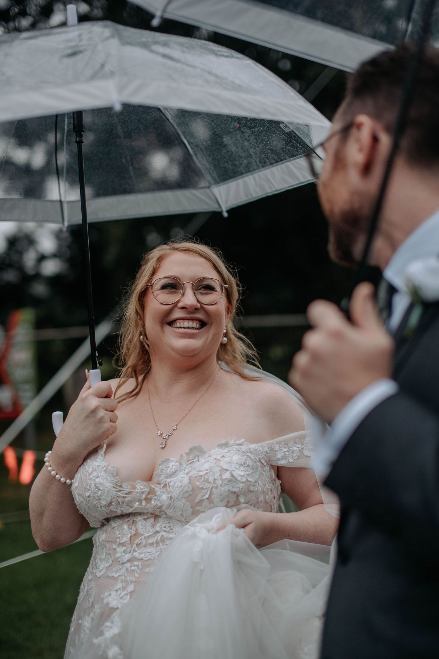 Couple sharing joyful moment under clear umbrellas during rainy outdoor wedding.